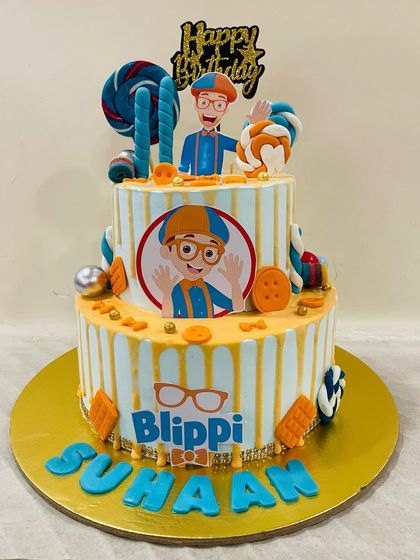 A cheerful two-tiered Blippi themed cake for a curious little explorer. The design is full of Blippi's signature orange and blue colors, buttons, and lollipops.