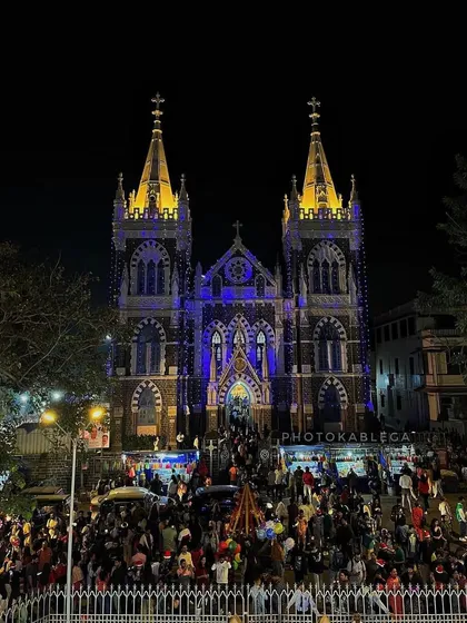 A similar stunning view of Mount Mary Basilica, capturing the Gothic architecture against the night sky, with crowds celebrating below.