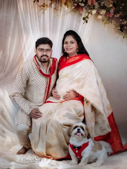 A lovely family portrait of an expecting couple and their pet, seated against an elegant, draped backdrop in our studio.