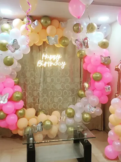 A beautiful first birthday decoration with a circular balloon arch in pink, peach, and gold. The design is accented with silver butterfly cutouts and a 'Happy Birthday' neon sign for a whimsical touch.