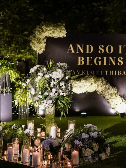 A personalized photo-op corner with the couple's hashtag. The backdrop is adorned with cascading white flowers, and the scene is lit by dozens of candles for a romantic glow.