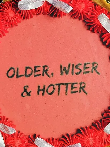 A top-down view of the 'Older, Wiser & Hotter' tart, showing the clean lettering and the vibrant contrast between the red topping and white ribbons. It's a fun and delicious way to celebrate another year.