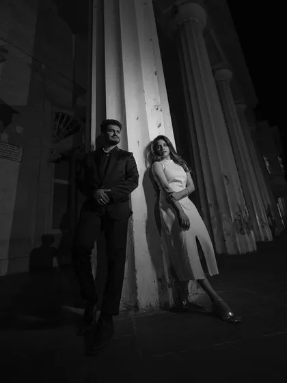 A stylish, high-contrast black and white portrait of a couple against architectural columns, creating a dramatic, editorial feel.