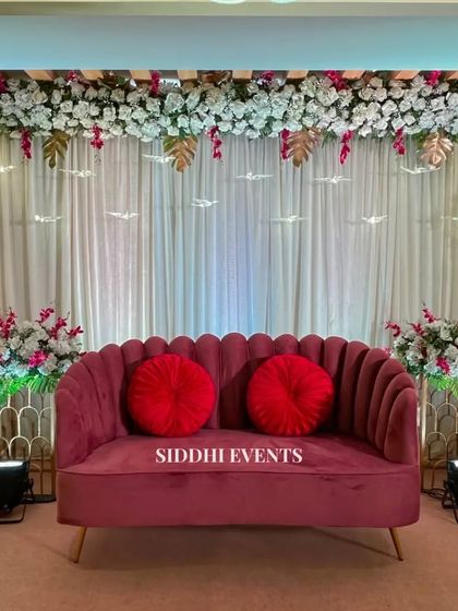 This elegant stage features a deep pink sofa with bold red cushions, set against a backdrop of white curtains and a canopy of white and pink flowers.