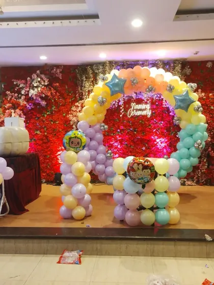 The stage setup for a naming ceremony with a colorful Cocomelon-themed balloon arch against a red floral wall.