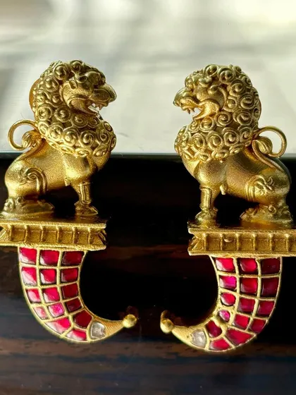 The iconic lion motif fusion earrings. These feature a detailed gold-plated lion stud with a red enamel horn-shaped drop, creating a bold and powerful statement piece.