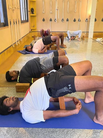 A group of tennis players practices a supported Setu Bandhasana (Bridge Pose) using wooden blocks. This restorative backbend helps to open the chest and hip flexors while relieving lower back tension, an essential recovery pose for athletes.