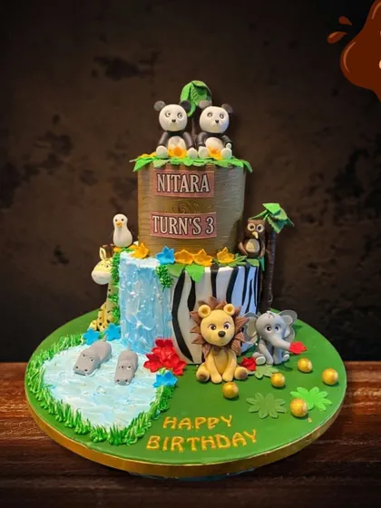 I designed this detailed jungle waterfall cake for a third birthday. It features two tiers with different animal prints and textures, a cascading blue waterfall, and a variety of cute fondant animals like pandas, hippos, and a lion.