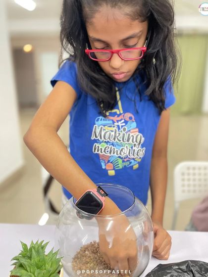 A young girl with pink glasses focuses on adding a layer of sand to her terrarium, a fun and tactile part of the process.