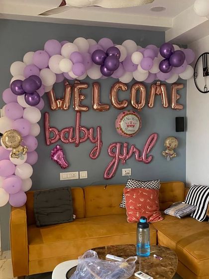 A 'Welcome Baby Girl' decoration in a living room. The setup includes a purple and white balloon arch and rose gold foil letters.