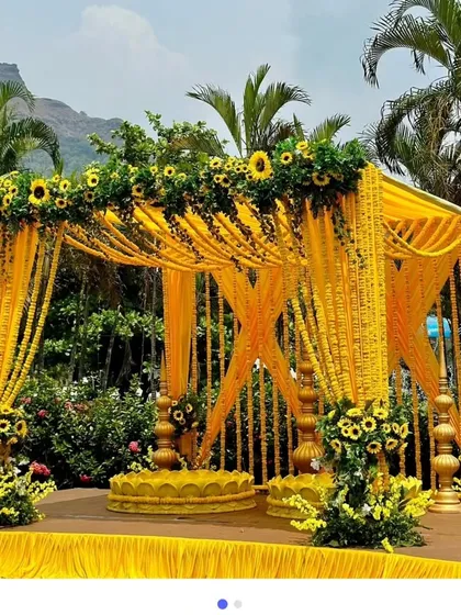 A bright and cheerful outdoor wedding mandap, perfect for a daytime ceremony. The entire structure is decorated with yellow drapes and strings of fresh marigolds, with sunflowers adding a beautiful touch to the canopy.