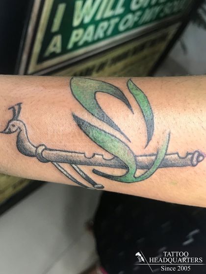 A custom tattoo design featuring a flute with the letter 'S' artistically integrated with green-colored feathers.