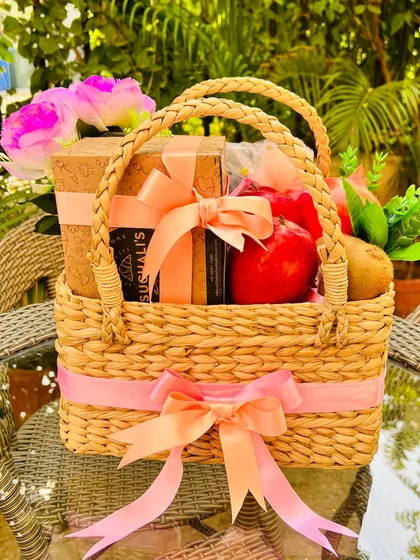 My modern interpretation of a classic fruit basket, combining health and indulgence. This handwoven cane basket features fresh, exotic fruits like apples and kiwis alongside a box of gourmet cookies, all tied with a beautiful peach and pink ribbon.