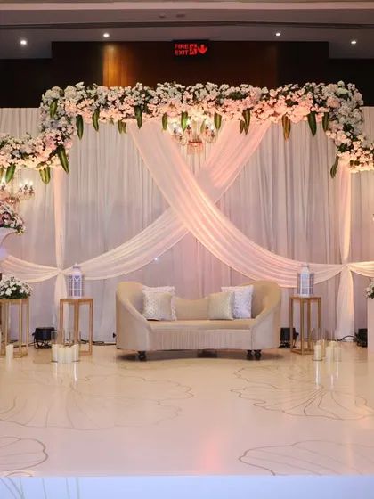 This elegant reception stage features a backdrop of soft, crisscrossing white drapes, framed by a beautiful arch of white and pink florals, creating a romantic and sophisticated look.