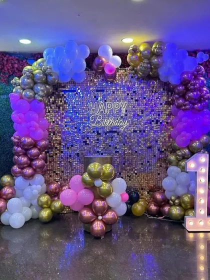 A glamorous first birthday setup with a gold sequin wall, a "1" marquee number, and a colorful balloon arch in pink, purple, and gold.