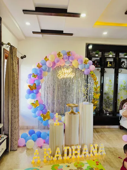 A clean shot of the pastel butterfly themed birthday setup without people, showcasing the full arrangement of the balloon arch, shimmer backdrop, pedestals, and marquee letters.