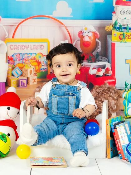 A happy baby boy sits on a miniature bed surrounded by all his favorite toys, looking directly at the camera with a big smile.