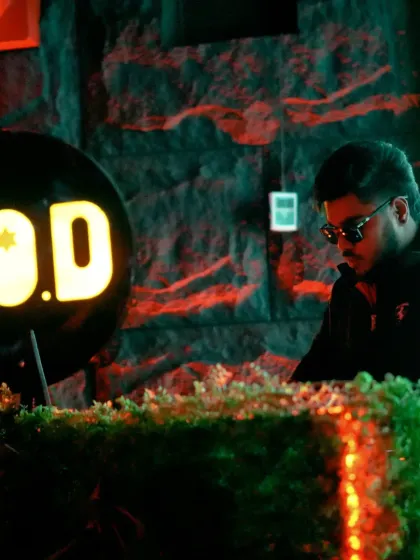A focused shot of me behind the decks at HOD, surrounded by lush greenery and the club's signature neon sign.
