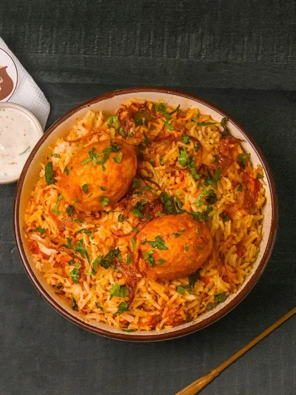 A delicious bowl of egg biryani, garnished with fresh herbs and served with a golden spoon. The dark background enhances the rich colors and makes the dish look very appealing.