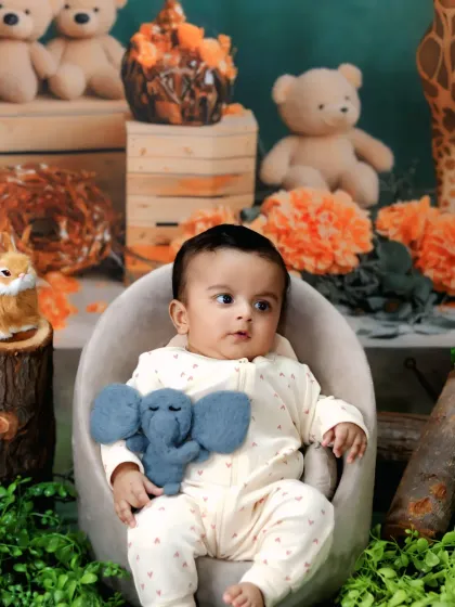 A calm and curious baby sits in a small armchair holding a toy elephant, surrounded by other plush animal friends.