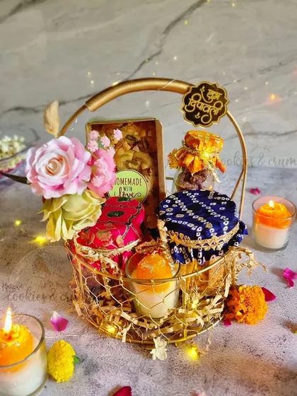 For a touch of elegance, I offer these beautiful golden baskets. This custom Diwali hamper includes assorted treat jars with traditional fabric tops, a box of homemade sweets, and a scented candle, all decorated with fresh flowers.