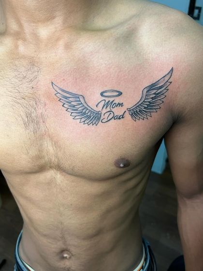 A classic "Mom Dad" tribute tattoo with angel wings and a halo. This chest piece is a popular way to honor parents.