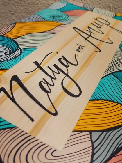 A close-up on the names "Natya and Anup," showing the smooth flow of the hand-painted calligraphy on the natural wood panel.