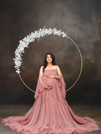 A lovely studio portrait of a mom-to-be in a pink gown, framed by a delicate floral hoop. The dark, textured background makes the subject and the floral element stand out beautifully.