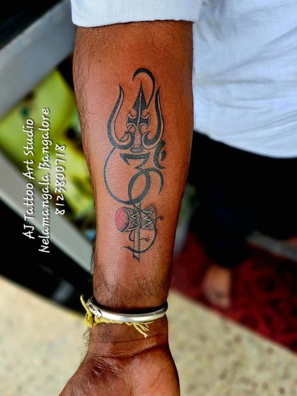 A powerful forearm tattoo combining the Trishul, Om symbol, and a Damaru with a splash of red. This is a classic and popular spiritual design.
