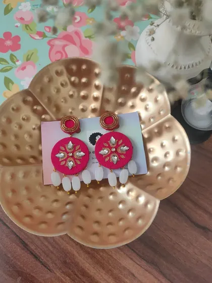 A close-up of my hand-painted fabric earrings. This pair features intricate white and gold floral patterns on a bright pink base, with delicate white bead drops for a festive look.