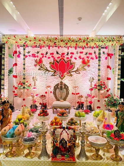 A grand and festive setup for a puberty ceremony. The backdrop features a large lotus motif made of flowers, surrounded by traditional brass lamps and a beautiful display of offerings.