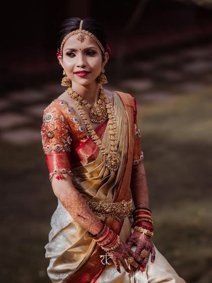 A portrait of a bride in her Muhurtham attire. The off-white and red silk saree is paired with a blouse featuring stunningly detailed embroidery on an orange base, creating a warm and radiant look.