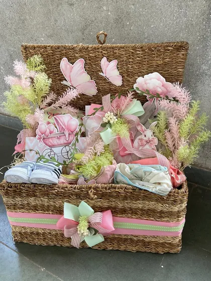 A charming wicker chest filled with pastel-colored baby gifts. The hamper is adorned with delicate butterfly and floral accents, creating a soft and dreamy "secret garden" theme.
