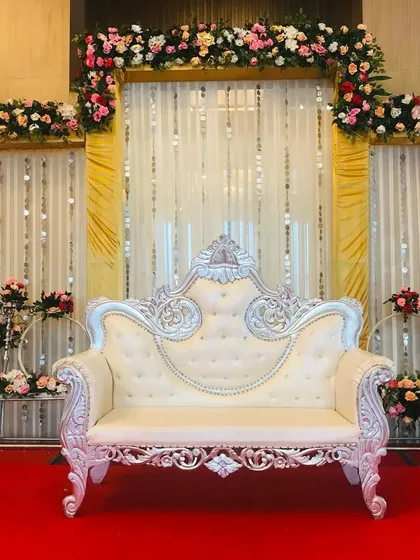 An elegant stage for a ring ceremony, featuring a silver-carved sofa against a backdrop of golden drapes and white floral arrangements.