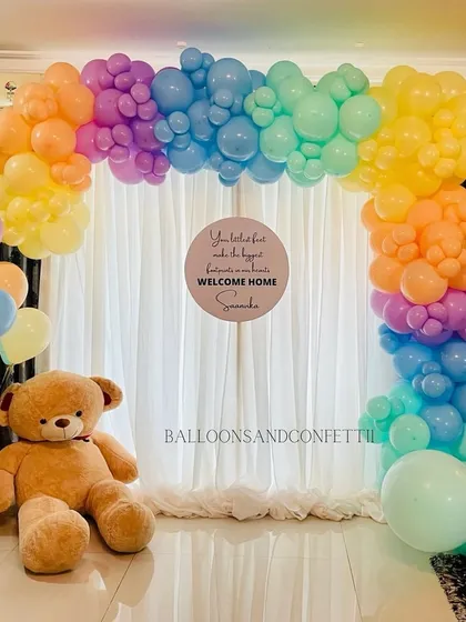 A vibrant rainbow-themed welcome home setup. The colorful balloon arch is paired with a giant teddy bear for a cheerful and heartwarming display.