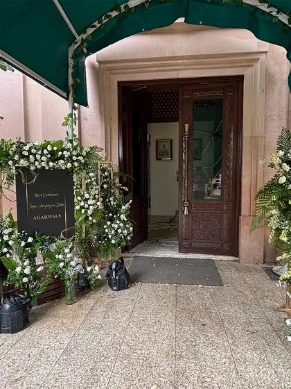 An elegant and understated welcome setup at the entrance of a classic venue. A simple black sign with gold lettering is beautifully framed by arrangements of fresh white roses and green ferns.