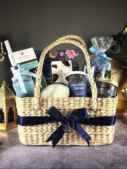 The 'Grand Galore' hamper is a basket filled with an assortment of treats in a cool blue theme, including a reed diffuser, coffee, and chocolates, tied with a navy blue ribbon.