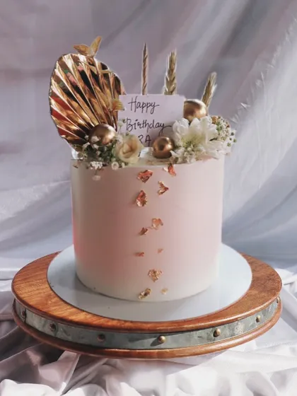 A lovely single-tier cake with a soft pink hue, decorated with a gold-painted palm spear, gold leaf, and delicate white flowers for a touch of boho glam.