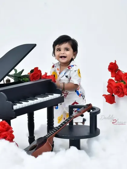 A charming studio session featuring a baby boy with a miniature grand piano. The set is decorated with red roses and fluffy clouds for a dramatic and adorable look.
