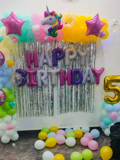 A whimsical unicorn-themed birthday decoration. This setup includes a silver fringe backdrop, a "Happy Birthday" balloon banner, a large unicorn foil balloon, and a colorful pastel balloon arch with star accents.