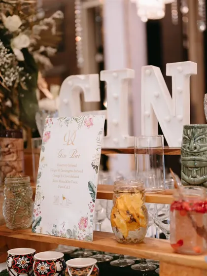 A rustic and romantic Gin Bar for a daytime wedding. The setup includes infused water, fresh fruit garnishes, and a floral menu, all arranged on a wooden display with quirky tiki mugs.