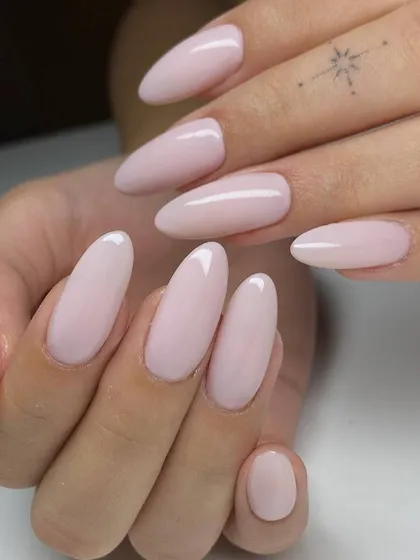 A beautiful, soft pink nude manicure on long, almond-shaped nails. The subtle star tattoo on the finger adds to the delicate and stylish aesthetic.