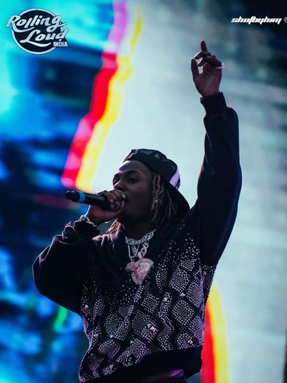 Rich the Kid hypes up the crowd at Rolling Loud India, his hand raised high. The colorful stage background and his iced-out jewelry are captured in sharp detail.