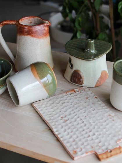 A charming set of student pieces including a pitcher, a house shaped lidded pot, and a textured platter, all in earthy green and white glazes.
