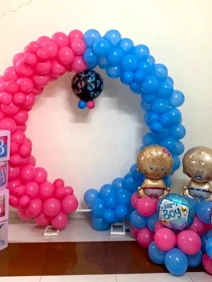 A gender reveal or baby shower decoration with a half-pink, half-blue balloon arch. The setup includes 'BABY' blocks and baby-shaped foil balloons.