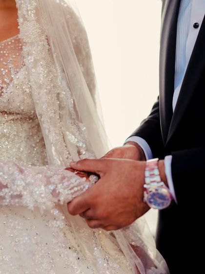 A close-up of the moment their hands meet. The bride's intricately beaded gown and the groom's classic tuxedo watch are details that tell the story of their personal style on their wedding day.