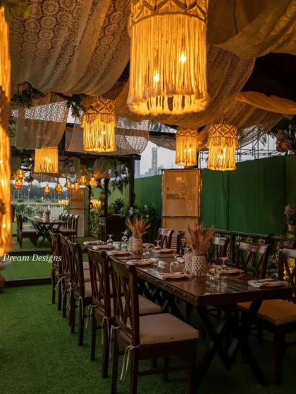 A detailed view of the rustic dining setup. The long wooden table is set under a canopy of draped lace and macrame lanterns, creating a warm and inviting atmosphere for an intimate dinner.