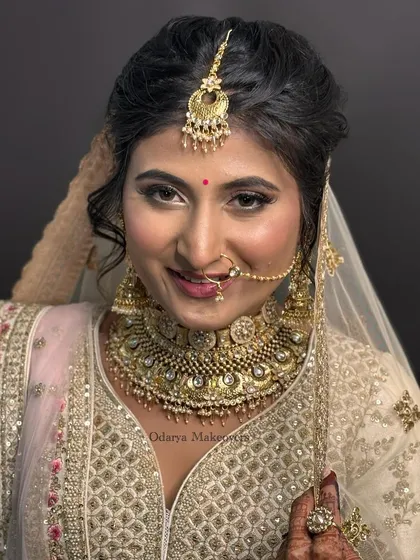 A stunning portrait of a Gujarati bride. The makeup features soft, shimmering eyeshadow and a warm lip color that beautifully complements her intricate gold jewellery and white lehenga.