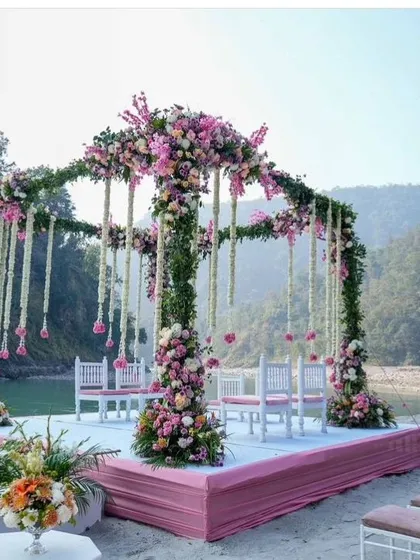 A beautiful riverside wedding mandap in Rishikesh. The structure is covered in lush greenery and pink flowers, with hanging floral strings adding a delicate touch.