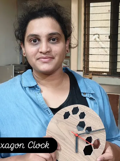 A student holding the modern hexagon clock she designed and built. This project involves learning to use a jigsaw for precise cuts and how to assemble the clock mechanism.